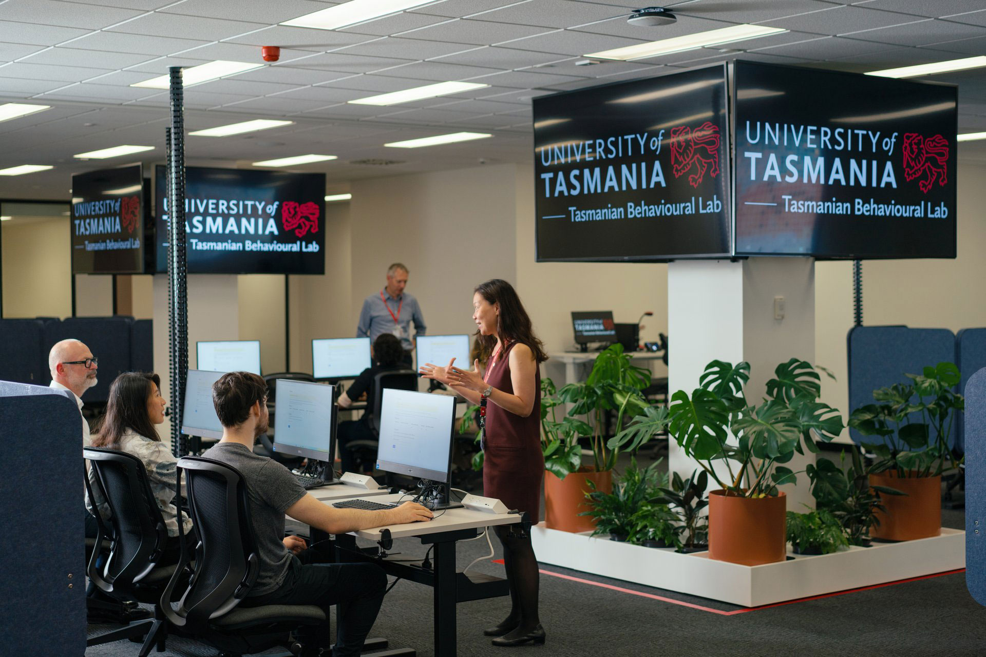Group of people in a collaborative office or lab with desks, computer monitors, and potted plants. Multiple screens display 'UNIVERSITY of TASMANIA Tasmanian Behavioural Lab.