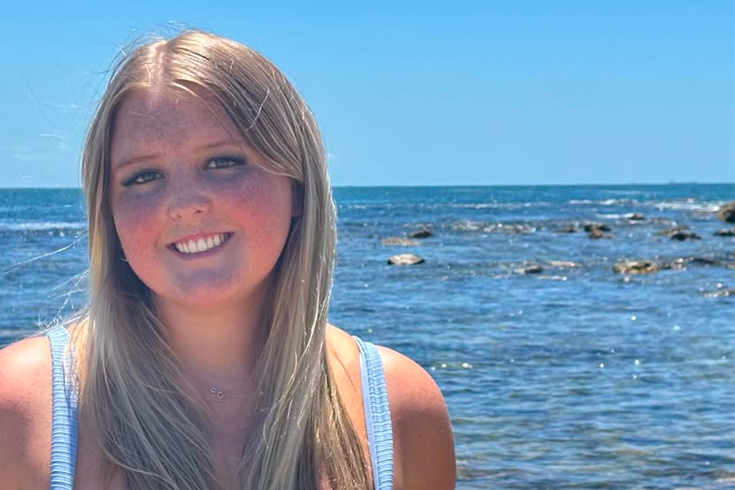 A young woman with long blonde hair stands close to the camera, with a serene blue sea, partially submerged rocks, and a clear blue sky in the background.